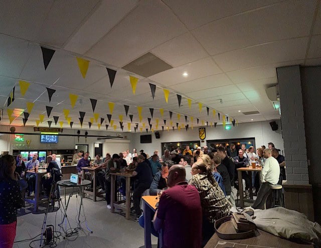 Drukke pubquizavond in een volle zaal met deelnemers aan hoge tafels en quizpresentatie voorin